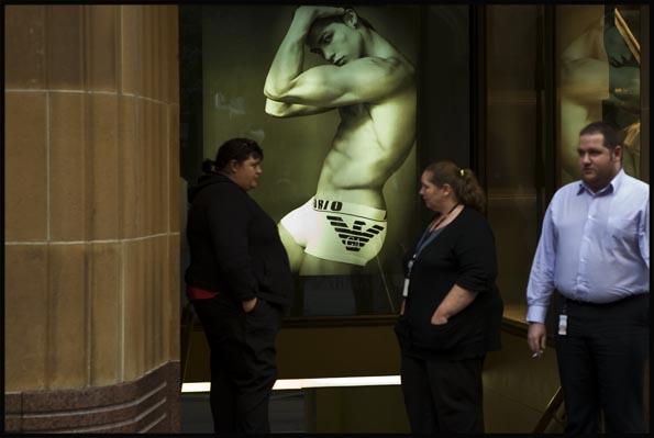 three overweight office workers having a cigarette break in front of an unlikely shop poster with a very fit male underwear model
